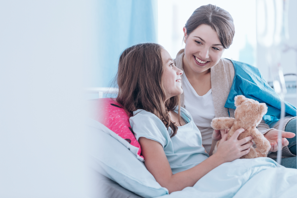 child laying in a hospital bed with a teddy bear with mom